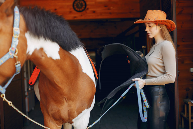 Mujer con sombrero poniendo la silla a un caballo para montarlo